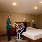 Woman assisting an elderly person using a patient transfer device lift in a bedroom setting.